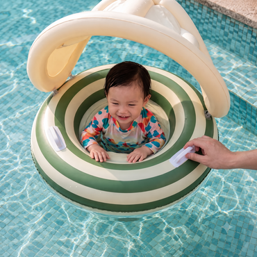 A baby wearing a colorful swimsuit sits inside a green and cream striped inflatable pool float with a canopy, held up by an adult hand in a tiled swimming pool.