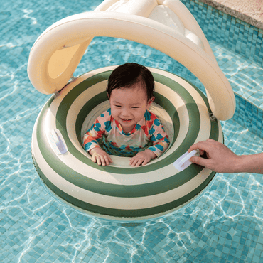 A baby wearing a colorful swimsuit sits inside a green and cream striped inflatable pool float with a canopy, held up by an adult hand in a tiled swimming pool.