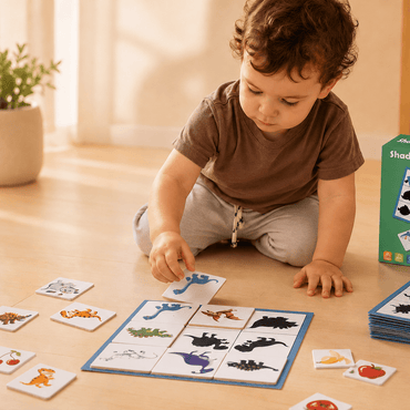 A toddler sits on a wooden floor playing with a shadow painting card game, with the green product box standing beside them.
