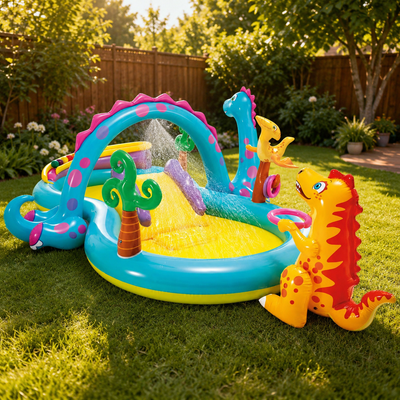 A colorful inflatable dinosaur-themed water play pool set on a green grass backyard. The pool features a turquoise base, a yellow water-filled center, pink polka-dot arches, green palm trees, and two dinosaur characters, one blue and one orange/yellow, positioned on either side.