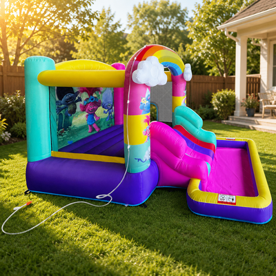 A colorful inflatable children's bounce house and slide set on a green backyard lawn. The play structure features a purple and teal bouncy area with a printed character scene, a rainbow archway, white cloud accents, a pink slide, and a matching pink playground pool.