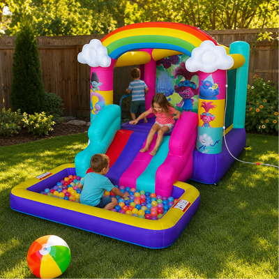 A colorful inflatable bounce house with a rainbow arch, cloud details, a slide, and a ball pit filled with children, set on a green backyard lawn.
