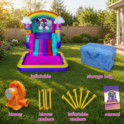 A colorful inflatable bounce house with slide is shown on a grassy backyard lawn, alongside its blue storage bag, orange blower, yellow anchors, and a small instruction manual. The bounce house has rainbow arches, cartoon character graphics, and purple, pink, yellow, blue, and green sections.