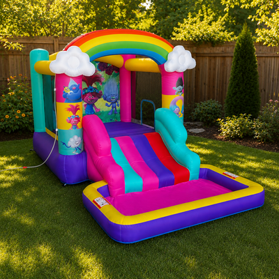 A colorful inflatable bounce house with a slide, set up on a green grass backyard with a wooden fence and trees in the background. The bounce house features a rainbow arch, Trolls-themed graphics, white clouds, and a pink, purple, blue, and teal slide.
